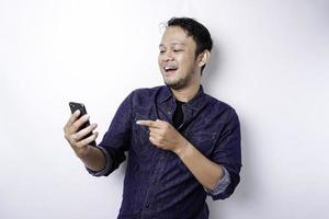 eccitato asiatico uomo indossare blu camicia sorridente mentre Tenere il suo Telefono, isolato di bianca sfondo foto