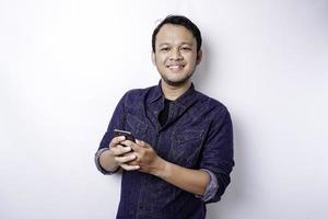 eccitato asiatico uomo indossare blu camicia sorridente mentre Tenere il suo Telefono, isolato di bianca sfondo foto