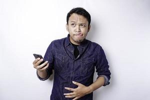 un' riflessivo asiatico uomo è Affamato e toccante il suo pancia mentre Tenere il suo Telefono pensiero che cosa per ordine, isolato di bianca sfondo foto