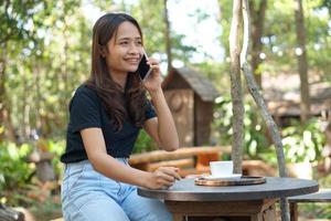 asiatico donna guardare a guadagni su sua Telefono a un' caffè negozio dove lei può partire per opera. in mezzo verde natura foto