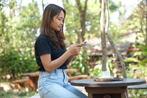 asiatico donna guardare a guadagni su sua Telefono a un' caffè negozio dove lei può partire per opera. in mezzo verde natura foto