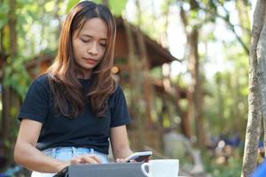 asiatico donna guardare a guadagni su sua Telefono a un' caffè negozio dove lei può partire per opera. in mezzo verde natura foto