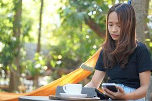 asiatico donna guardare a guadagni su sua Telefono a un' caffè negozio dove lei può partire per opera. in mezzo verde natura foto