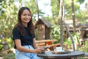 asiatico donna guardare a guadagni su sua Telefono a un' caffè negozio dove lei può partire per opera. in mezzo verde natura foto