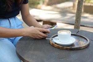 asiatico donna guardare a guadagni su sua Telefono a un' caffè negozio dove lei può partire per opera. in mezzo verde natura foto