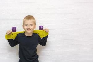 biondo ragazzo Tenere un' skateboard dietro a il suo le spalle, sorridente, contro il sfondo di un' bianca parete con copyspace, adolescenti attività foto