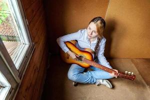 giovane fricchettone donna seduta su pavimento e giocando chitarra a casa. adolescente ragazza apprendimento per giocare canzone e scrittura musica nel sua camera. passatempo, stile di vita, relax, strumento, tempo libero, formazione scolastica concetto. foto