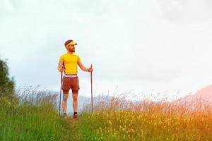 uomo con barba sembra lontano mentre a piedi su il sentiero nel il mezzo di montagna prati con bastone foto