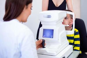 femmina oculista utilizzando macchina per controllo occhio vista nel clinica. poco ragazzo guardare a attrezzatura e medico analisi occhio allievo nel ottico negozio. concetto di occhio cura e Salute. foto