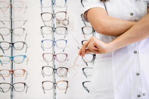 femmina oculista con attraversato braccia nel clinica. ritagliata Immagine di attraente giovane femmina medico nel oftalmologia clinica. medico oculista è in piedi vicino scaffali con diverso occhiali. foto