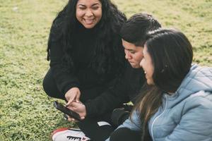 gruppo di latinos ridendo seduta su il terra nel un' parco con un' smartphone foto