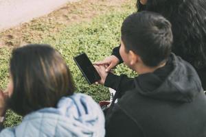 Visualizza a partire dal il indietro gruppo di ridendo latinos seduta su il terra nel un' parco con un' smartphone foto