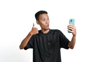 asiatico uomo indossare nero formazione maglietta, con un' contento eccitato gesto mentre guardare a il smartphone schermo. isolato di bianca sfondo foto