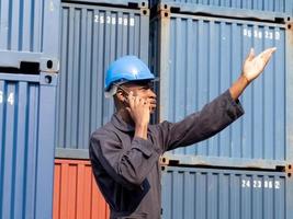 Sud africano nero pelle americano persona blu elmetto protettivo casco sicurezza uniforme persone puntamento dito allegro parlare smartphone mobile comunicazione opera lavoro occupazione attività commerciale importare esportare logistica foto