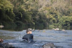 asiatico ragazzi siamo la spesa loro tempi liberi di immersione, nuoto, lancio rocce e attraente pesce nel il fiume insieme felicemente. passatempo e felicità di bambini concetto. foto