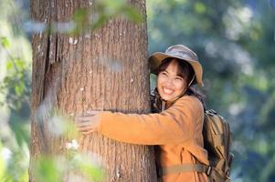 giovane turista donna nel un' giallo cappotto abbracciare un' albero nel il foresta di eco amore guardare su a il Treetops giovane asiatico donna l'esame un' grande ecologico albero foto