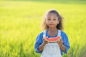 nero spellato carino poco ragazza mangiare anguria all'aperto verde riso campo fondale africano bambino mangiare anguria foto