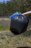 mano detiene contro un' foresta pieno di spazzatura un' grande nero plastica Borsa, un' estate giorno foto