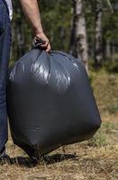 dell'uomo mano detiene contro un' foresta pieno di spazzatura un' grande nero plastica Borsa foto