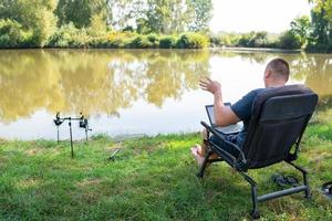 un' uomo si siede nel un' sedia vicino il autunno lago e lavori su un' il computer portatile libero professionista campeggio. robot su di il ufficio, telefonato con colleghi. foto