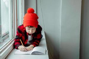 ragazzo fa compiti a casa. il ragazzo bugie su il davanzale e scrive nel un' taccuino. scolaro fare compiti a casa di il finestra. casa scolarizzazione. foto