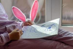 un' scolara ragazza con coniglio orecchie si siede su il finestra e disegna un' coniglio nel sua album, preparazione per Pasqua foto