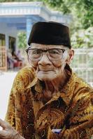 un' ritratto di un vecchio asiatico uomo indossare batik camicia e bicchieri foto