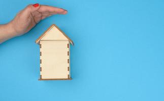 femmina mano al di sopra di di legno miniatura modello Casa su blu sfondo. vero tenuta assicurazione concetto, ambientale protezione foto