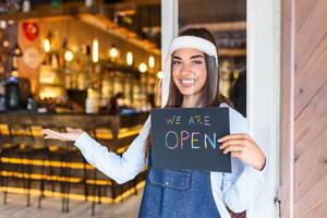 piccolo attività commerciale proprietario sorridente mentre Tenere il cartello per il riapertura di il posto dopo il quarantena dovuto per covid19. donna con viso scudo Tenere cartello noi siamo aprire, supporto Locale attività commerciale. foto