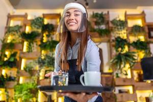femmina cameriera indossare viso scudo, visiera servi il caffè nel ristorante durante coronavirus pandemia che rappresentano nuovo normale concetto foto