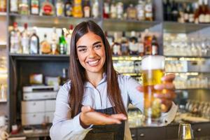 bellissimo sorridente femmina barista servendo un' bozza birra a il bar contatore , scaffali pieno di bottiglie con alcool su il sfondo foto