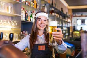 barista con viso scudo, covid-19 protezione, servendo un' bozza birra a il bar contatore durante coronavirus pandemia, mostrando ok segno, mensole pieno di bottiglie con alcool su il sfondo foto