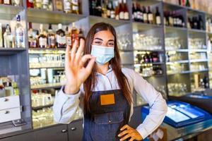 contento giovane donna indossare protettivo viso maschera nel il ristorante durante coronavirus pandemia, barista mostrando ok cartello. scaffali pieno di bottiglie con alcool su il sfondo foto