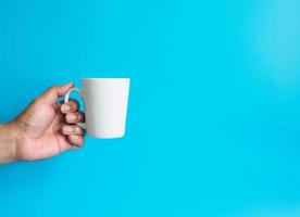 uomo mano Tenere un' bianca caffè tazza, pieno con caldo nero caffè Americano, no zucchero no latte, pronto per bere, rinfrescante. aroma sveglio fresco per opera posto su un' blu isolato sfondo foto