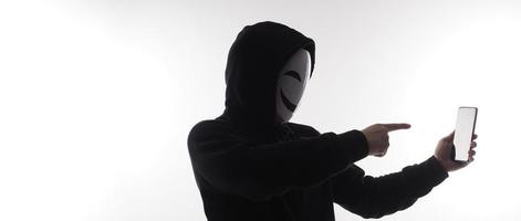 pirata anonimo e viso maschera con smartphone nel mano. uomo nel nero cappuccio camicia Tenere e utilizzando mobile Telefono su bianca sfondo. rappresentare informatica crimine dati pirateria o furto personale dati concetto foto