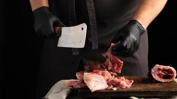 capocuoco nel nero latice guanti detiene un' grande coltello e tagli in pezzi crudo coniglio carne su un' Marrone di legno taglio tavola foto