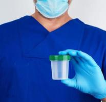 medico nel blu uniforme e latice guanti è Tenere un vuoto plastica contenitore per assunzione urina campioni foto