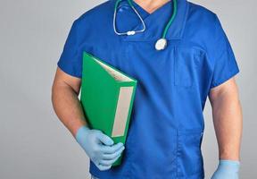 medico nel blu uniforme, latice sterile guanti detiene nel il suo mano un' verde cartella foto