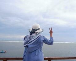 indietro di il donna nel il cappello su il tropicale spiaggia chi è guardare su a il cielo e il mare, mostrando pace gesto. verticale. mare sfondo foto