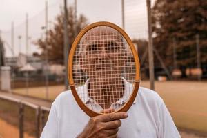 ritratto di anziano tennis giocatore vestito nel abbigliamento sportivo in posa con racchetta su viso su un' argilla tennis Tribunale foto