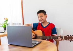 il storia di un' ragazzo Guardando un' taccuino computer mentre preparazione per pratica giocando chitarra a casa. ragazzi prendere classico chitarra Lezioni in linea. foto