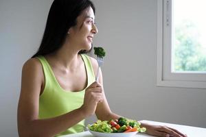 un' contento asiatico donna con un' salutare insalata piatto, Tenere forchetta con un' grande broccoli. cibo per bene forma foto