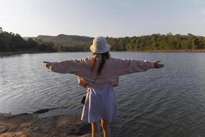 il indietro di un' giovane donna indossare un' alla moda vestito indossare un' cappello in piedi con sua mani disteso nel il luce del sole sensazione nostalgico per il bellissimo natura foto