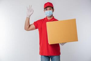 Immagine di un' consapevole giovane consegna uomo nel rosso berretto vuoto maglietta uniforme viso maschera guanti in piedi con vuoto Marrone cartone scatola isolato su leggero grigio sfondo studio foto