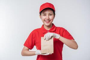 Immagine di un' contento giovane consegna uomo nel rosso berretto vuoto maglietta uniforme in piedi con vuoto Marrone mestiere carta pacchetto isolato su leggero grigio sfondo studio foto