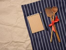 blu dello chef grembiule, di legno cucchiai e taccuino con vuoto Marrone lenzuola foto