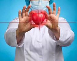medico nel un' bianca medico cappotto Tenere un' rosso cuore. cardiovascolare malattia concetto, presto diagnosi. blu sfondo foto