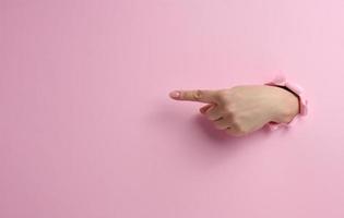 femmina mano bastoni su di un' strappato buco nel un' rosa carta sfondo, parte di il corpo punti con il indice dito per il lato foto