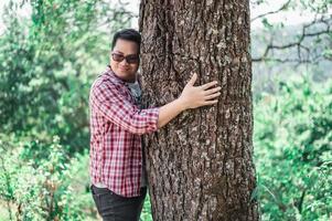 ritratto di contento asiatico uomo abbracciare un' albero nel foresta foto
