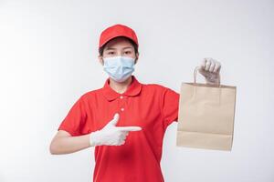 Immagine di un' contento giovane consegna uomo nel rosso berretto vuoto maglietta uniforme viso maschera guanti in piedi con vuoto Marrone mestiere carta pacchetto isolato su leggero grigio sfondo studio foto